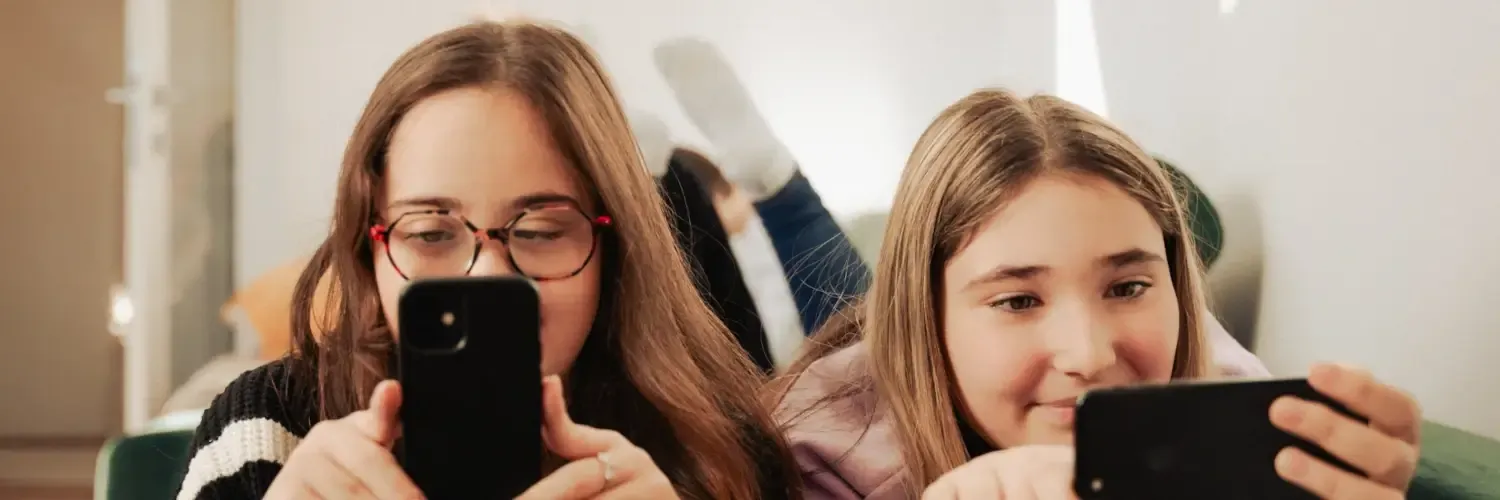 Isabell and Stella are lying next to each other on a dark green couch on their stomachs, looking at their smartphones.